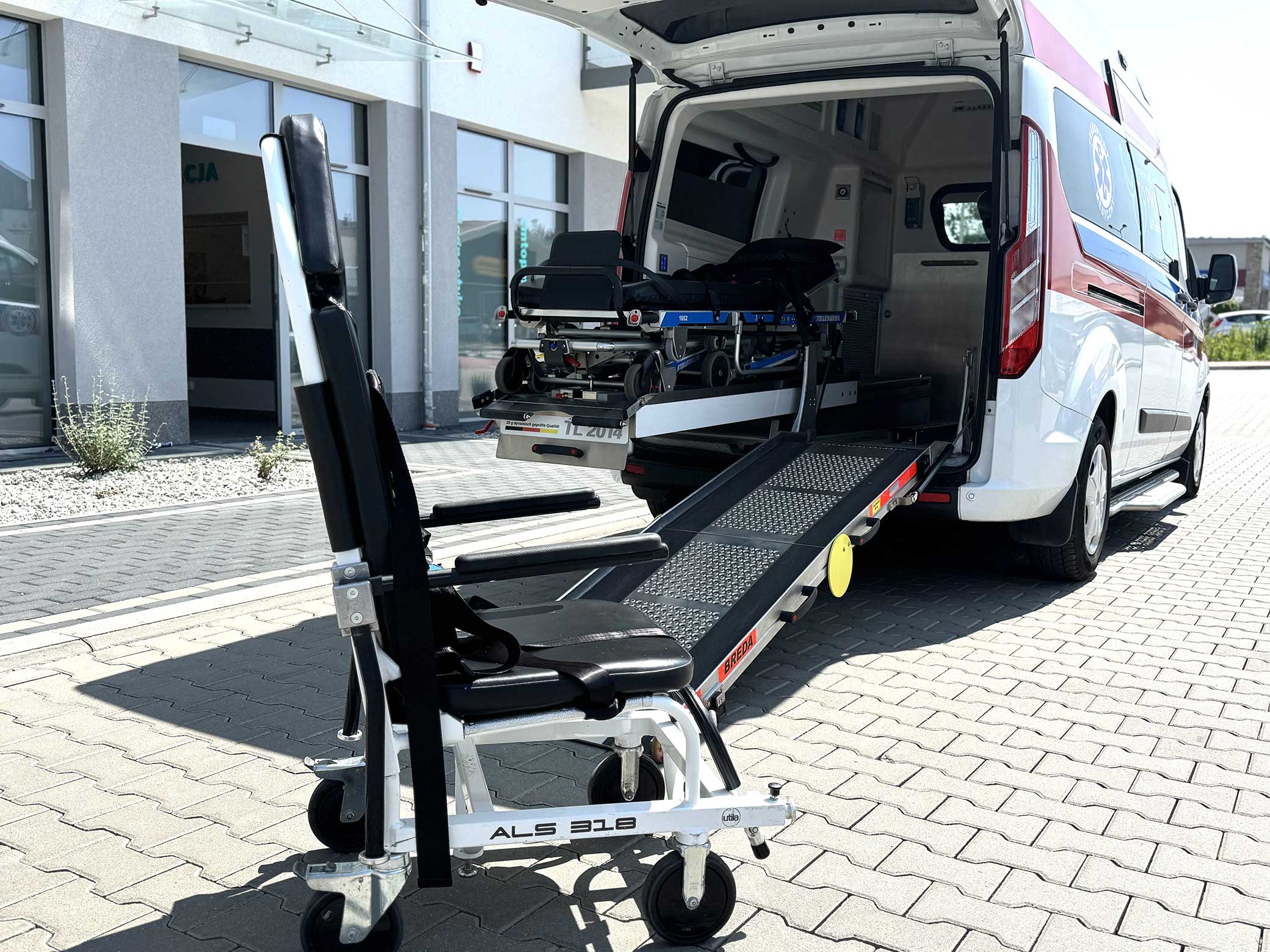 wózek dla pacjenta stoi przed karetką z otwartymi z tyłu drzwiami, wysuniętymi noszami i podjazdem